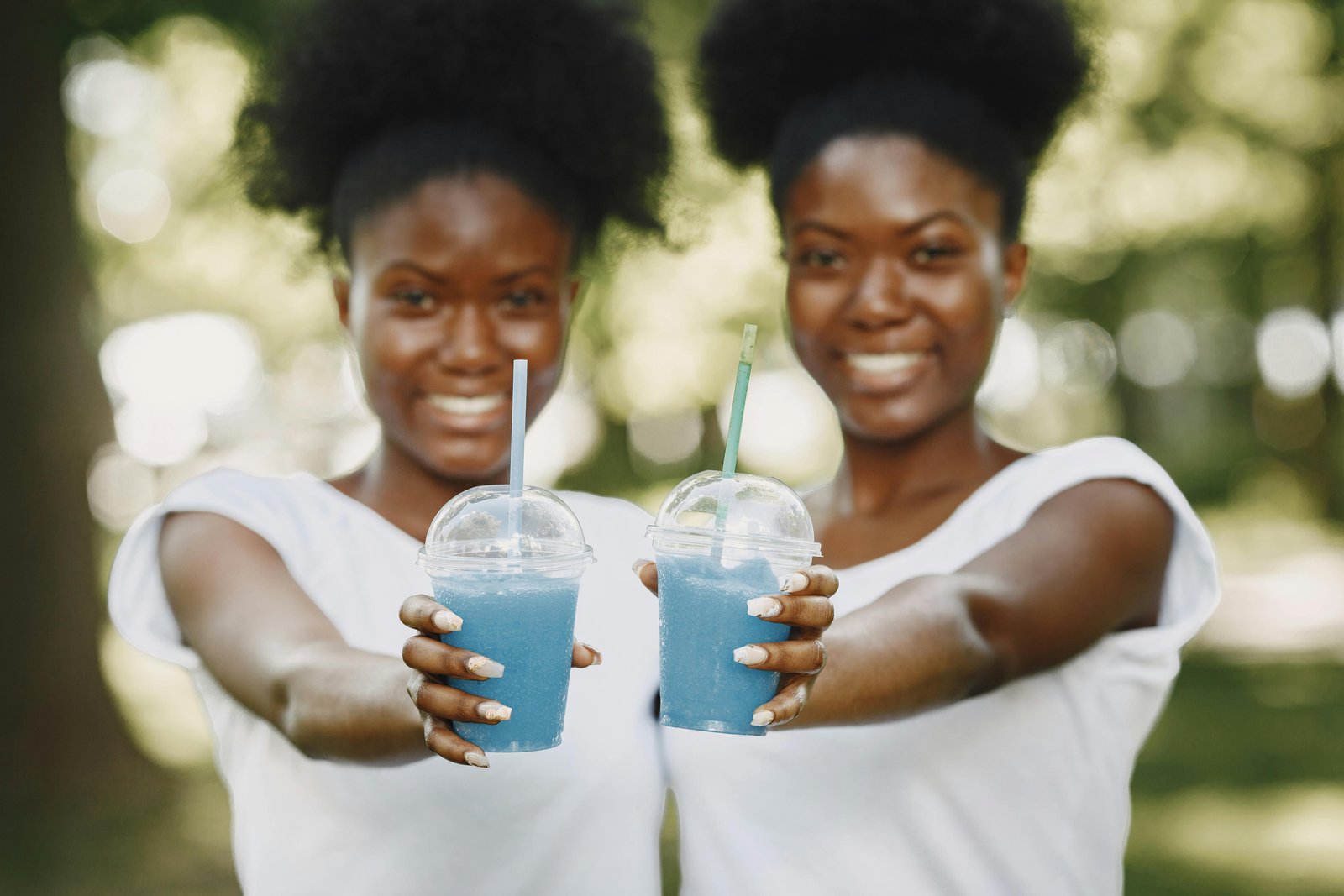 Twin sisters enjoying blue smoothies in a park, showcasing smiles and summer vibes.