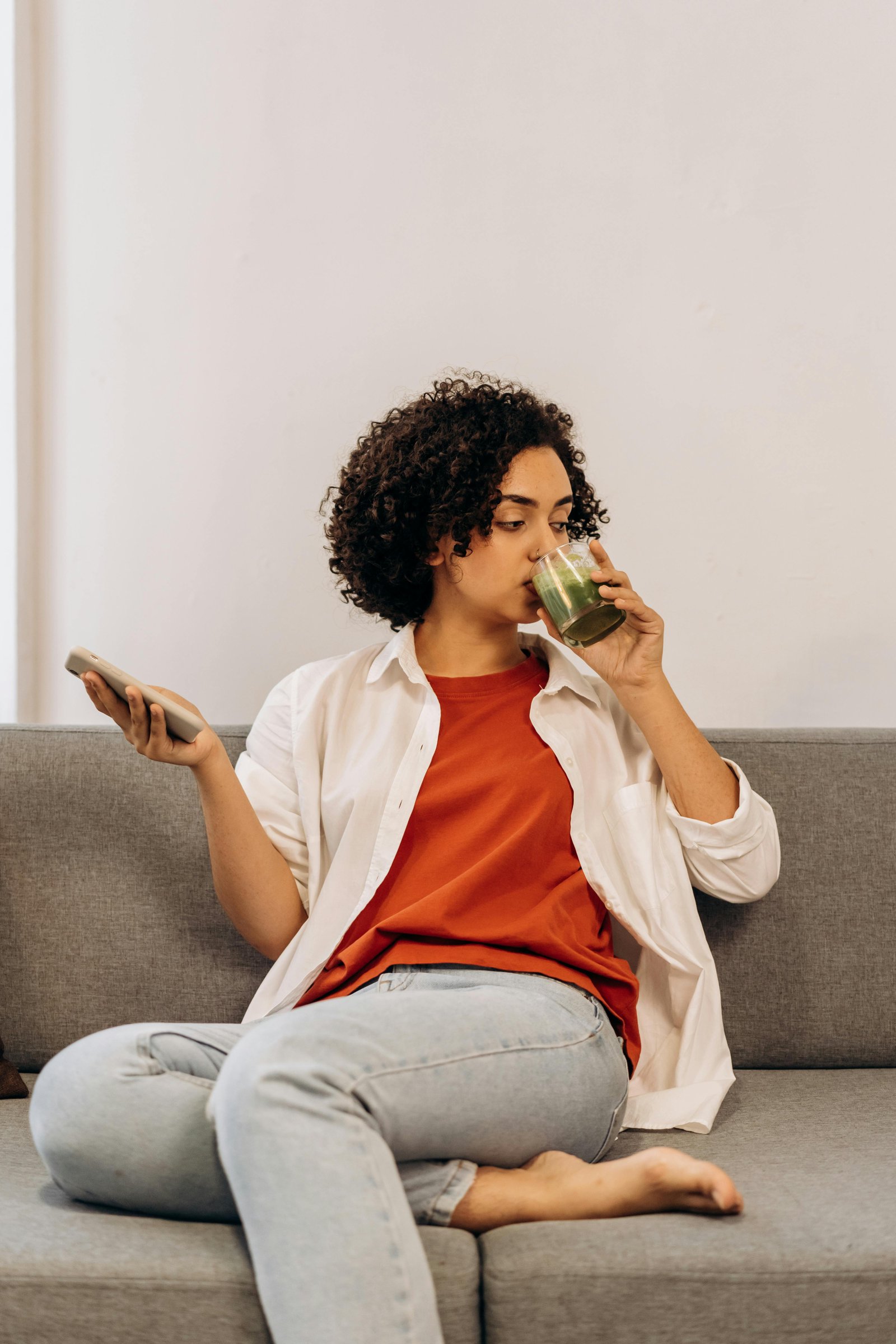 A woman comfortably seated indoors sipping on a nutritious green smoothie while holding a smartphone.