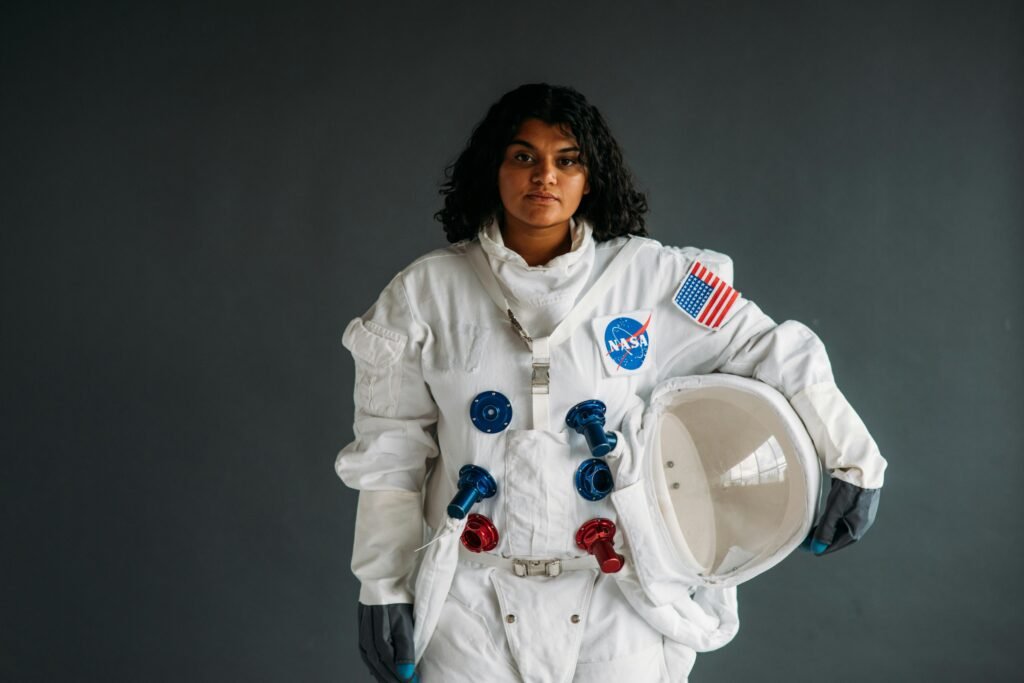 Woman dressed as astronaut holding helmet in NASA suit against grey background.