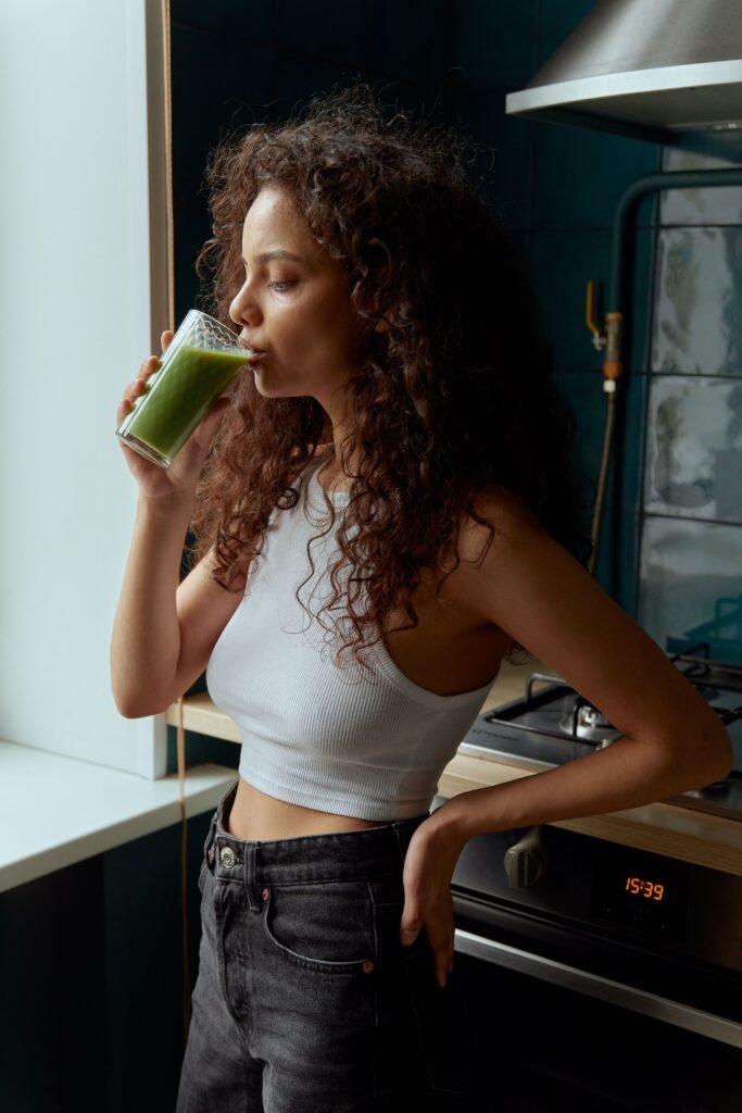 Young woman with curly hair drinks green smoothie indoors. Emphasizes healthy lifestyle.